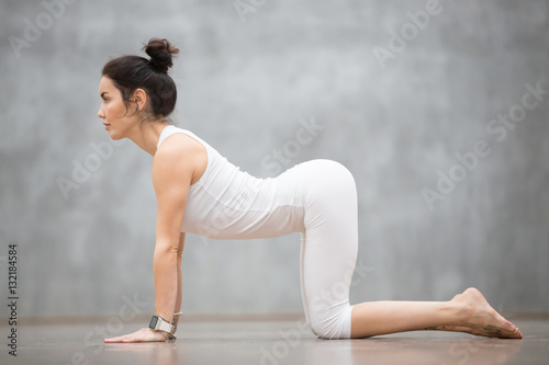 Beautiful young woman with tattoo on her foot meaning Wild cat working out against grey wall, doing yoga or pilates exercise. Cow, Bitilasana, asana paired with Cat Pose on the exhale. Full length