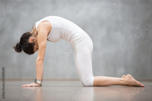 Beautiful young woman with tattoo on her foot meaning Wild cat working out against grey wall, doing yoga or pilates exercise. Cat, Marjaryasana, asana paired with Cow Pose on the inhale. Full length