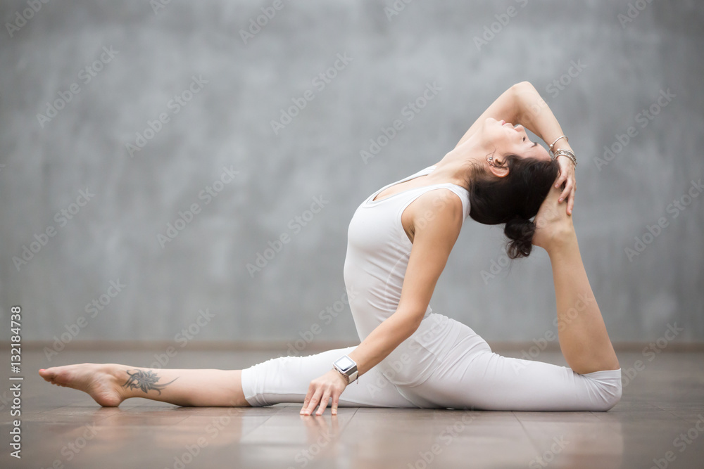 Young pretty woman wearing white sportswear set and smartwatch working out against grey wall, doing yoga or pilates exercise. Variation of monkey god, splits, hanumanasana with backbend. Full length