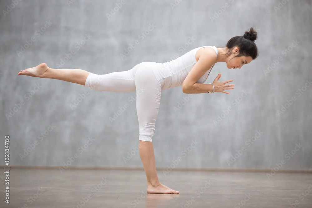 Side view portrait of beautiful young woman wearing white tank top working out against grey wall, doing yoga or pilates exercise. Standing in Warrior three pose, Virabhadrasana 3. Full length