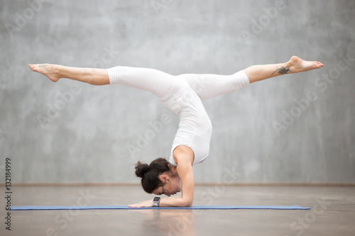 Portrait of beautiful young woman wearing white sportswear working out against grey wall, doing yoga or pilates exercise. Handstand with splits, variation of Pincha Mayurasana. Full length shot