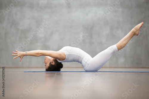 Side view portrait of beautiful young woman wearing white sportswear working out in fitness center against grey wall  doing yoga or pilates exercise. Variation of Matsyasana  Fish pose. Full length