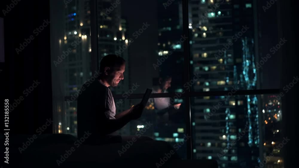 Young man browsing web on tablet computer sitting on bed at home at night
Young man browsing web on tablet computer sitting on bed at home at night