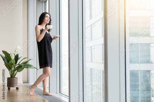 Full length view of young woman standing near window enjoying coffee, looking at city after waking up at morning, wearing black silk nightwear, resting from the business, waiting for good weather