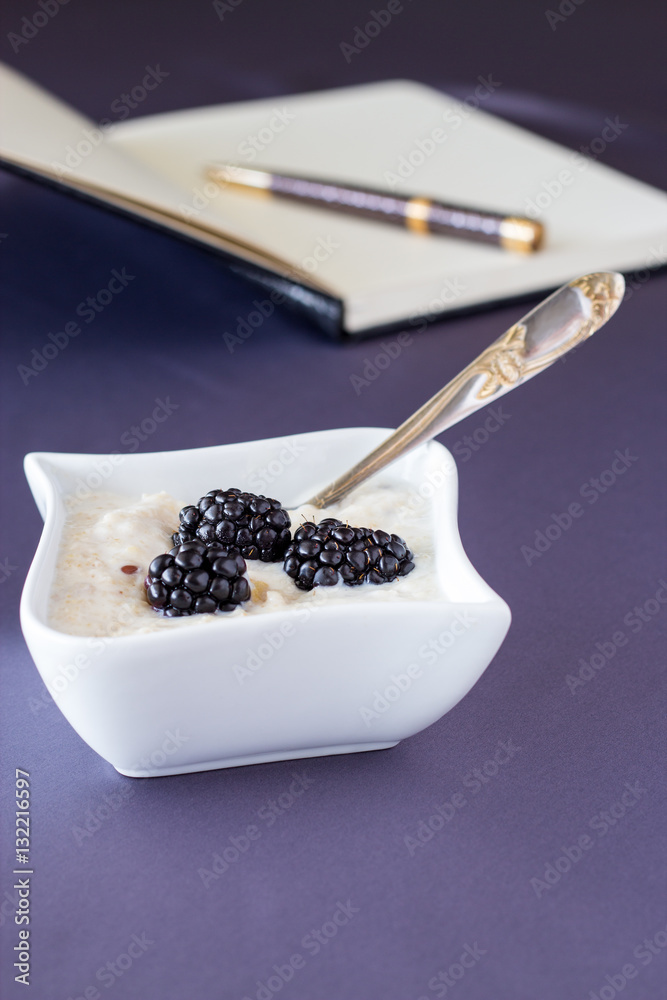 Healthy oatmeal with dried fruit and blackberries in a white bowl of irregular shape, notebook exercise book and pen in the background, neutral background Healthy oatmeal with dried fruit and blackberries in a white bowl of irregular shape, notebook exercise book and pen in the background, neutral background