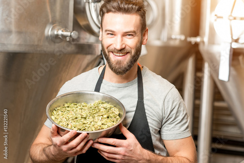 Portrait of a handsome brewer in uniform with hop in the bowl at the manufacturing with metal tanks