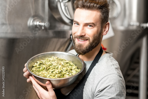 Portrait of a handsome brewer in uniform with hop in the bowl at the manufacturing with metal tanks