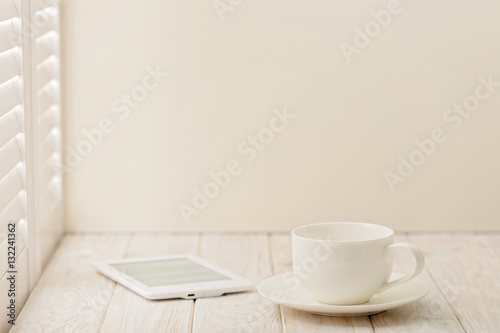 E-book and a cup on a light wooden background near a window with