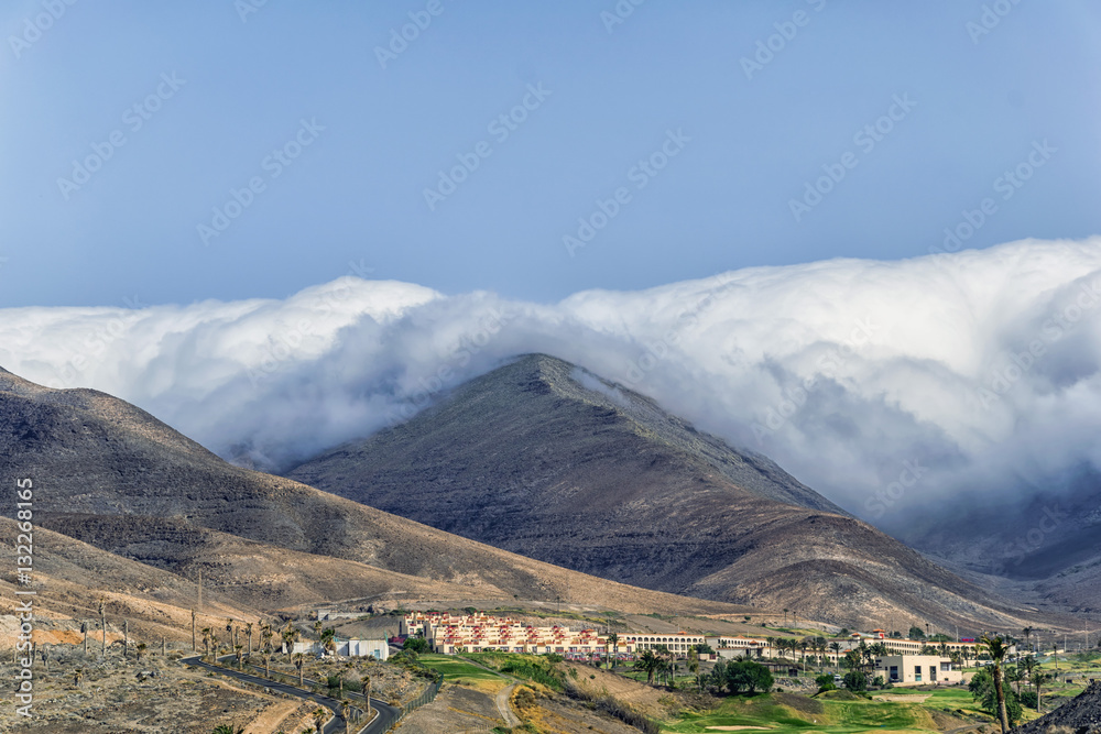 Einzigartige Landschaft auf Fuerteventura, Kanarische Inseln, Spanien mit einem Dorf vor der trockenen, vulkanischen Berge bedeckt in weißen dicken Wolken.