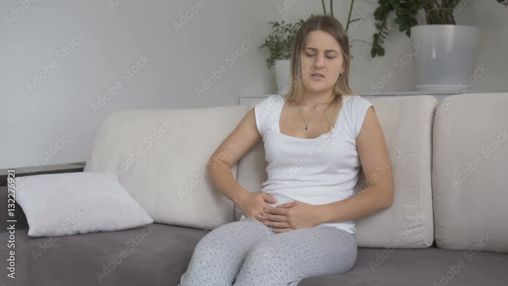 Young woman with menstrual pain sitting on sofa and holding hands on stomach Young woman with menstrual pain sitting on sofa and holding hands on stomach