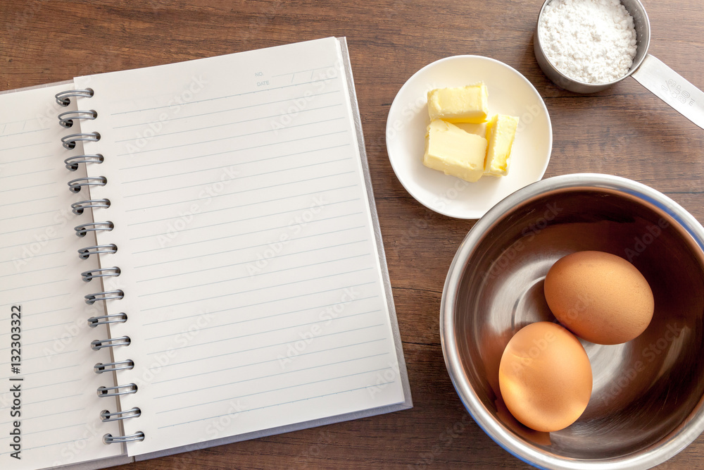 Blank notebook with eggs, butter and flour in measure cup on the wooden table