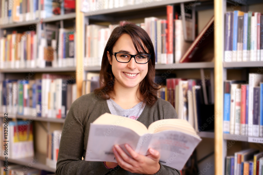 long brown hair girl with black glasses holding book and smiling. portrait of female student in a library with bookshelf on background long brown hair girl with black glasses holding book and smiling. portrait of female student in a library with bookshelf on background