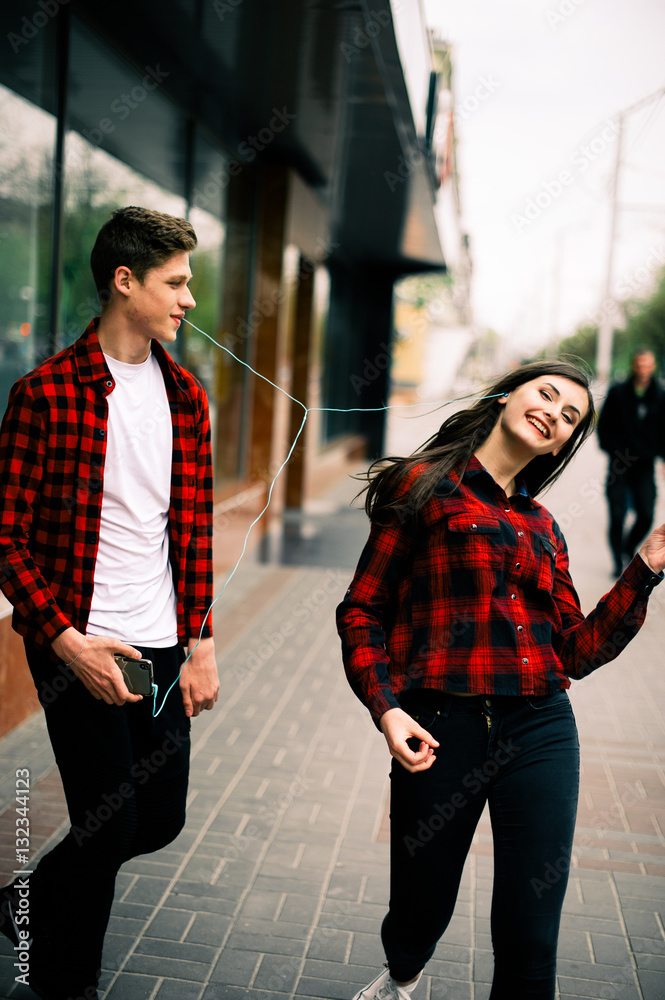 Naklejka premium Two happy trendy teenage friends walking and dancing in the city, listening to the music with headphones, talking each other and smiling. Lifestyle, friendship and urban life concepts.
