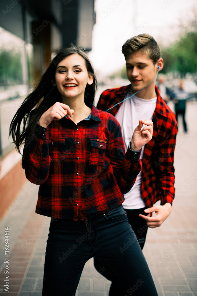 Two happy trendy teenage friends walking and dancing in the city, listening to the music with headphones, talking each other and smiling. Lifestyle, friendship and urban life concepts.