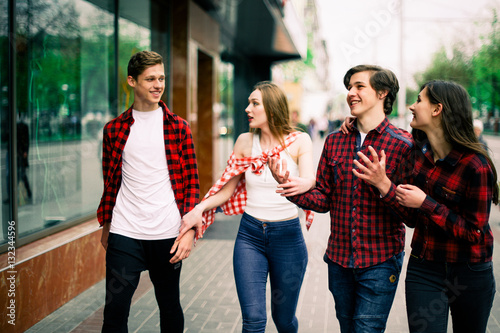 Four happy trendy teenage friends walking in the city, talking each other and smiling. Lifestyle, friendship and urban life concepts.