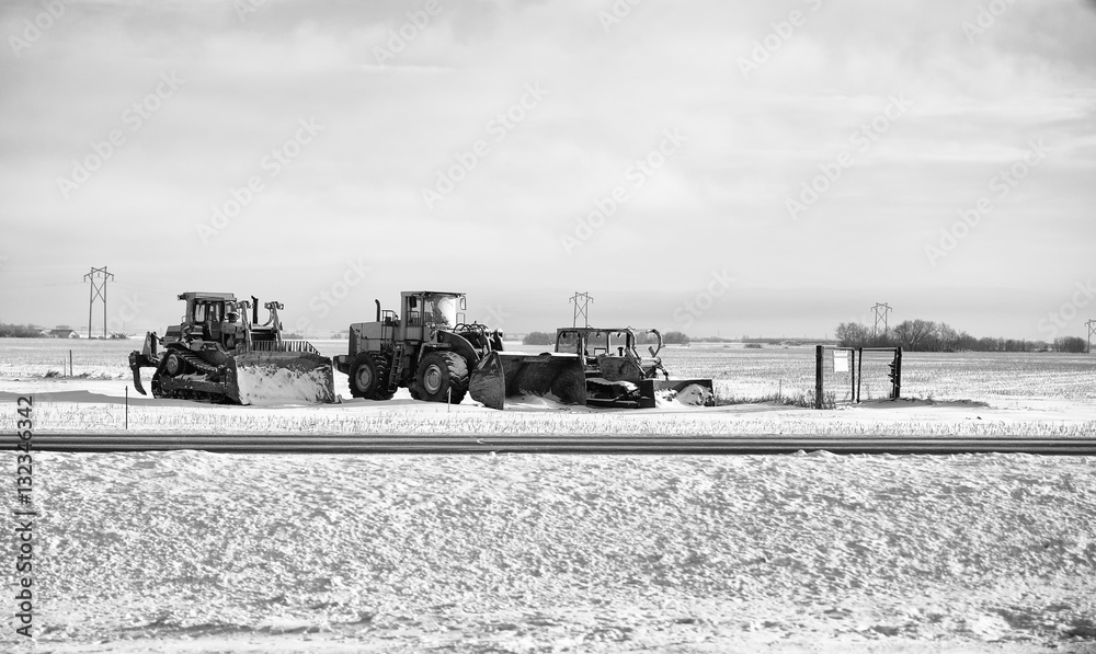 Three industrial snow removal equipment parked beside a highway on a field in rural black and white countryside winter landscape