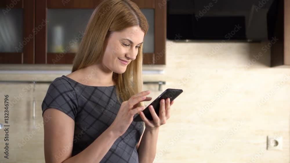 Beautiful young woman with a phablet standing in kitchen. Browsing Internet and smiling. Beautiful young woman with a phablet standing in kitchen. Browsing Internet and smiling.