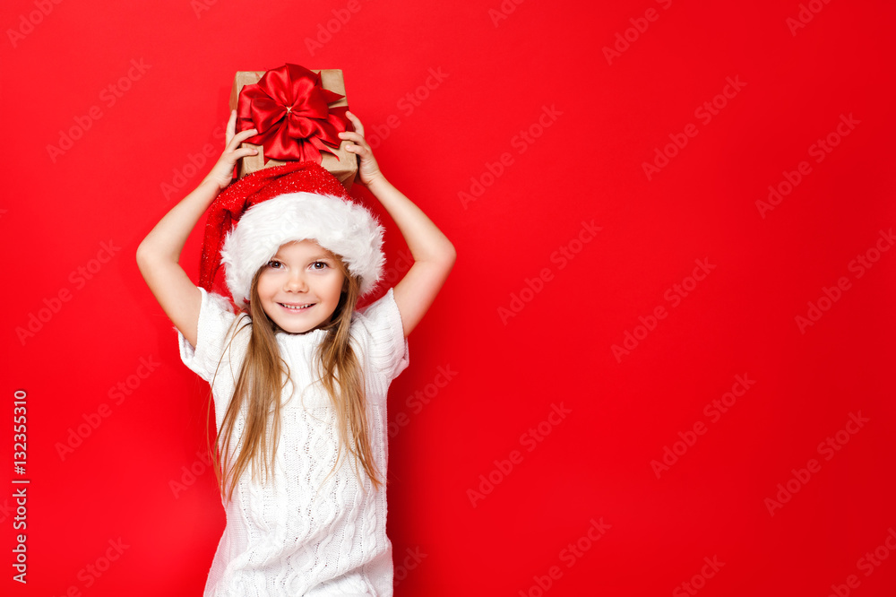 Happy girl in Christmas caps with gift wrapped in craft paper and red ribbons on red background. Christmas. Happy family. Happy girl in Christmas caps with gift wrapped in craft paper and red ribbons on red background. Christmas. Happy family.