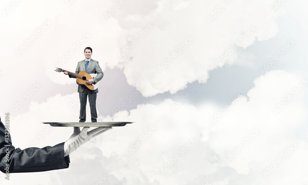 Businessman on metal tray playing acoustic guitar against blue sky background Businessman on metal tray playing acoustic guitar against blue sky background