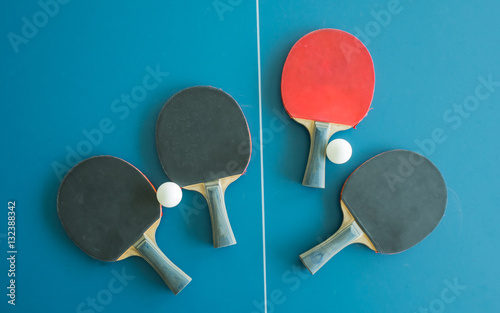 A red table tennis bat surrounded by three black table tennis bats and balls on a table with white vertical line. Concept of uniqueness, outstanding.