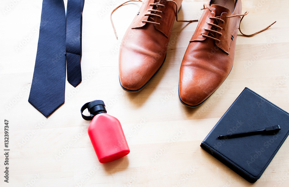 A men accessories laying down on wooden table, black necktie, leather brown shoe, red perfume bottle,black electronic reader