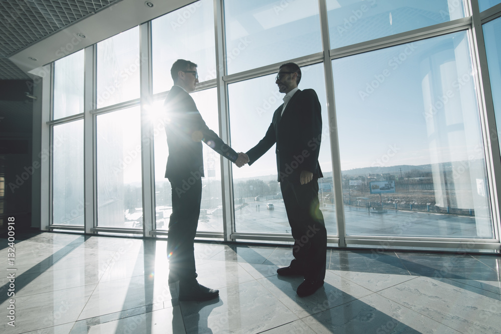 Full length side view of businessmen shaking hands in against panoramic windows Full length side view of businessmen shaking hands in against panoramic windows