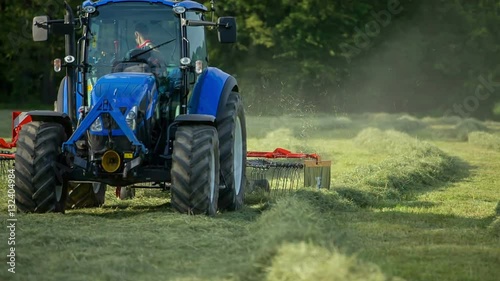 We can see a young farmer sitting in a blue tractor and he is raking hay with the help of a big machinery that is connected to his tractor. It is a nice summer day.
