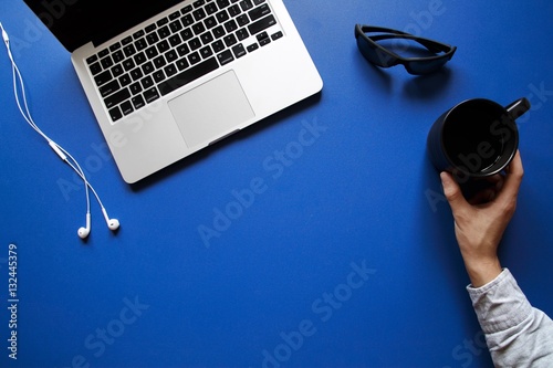 Hand placing mug next to laptop, sunglasses and earbuds on blue desk