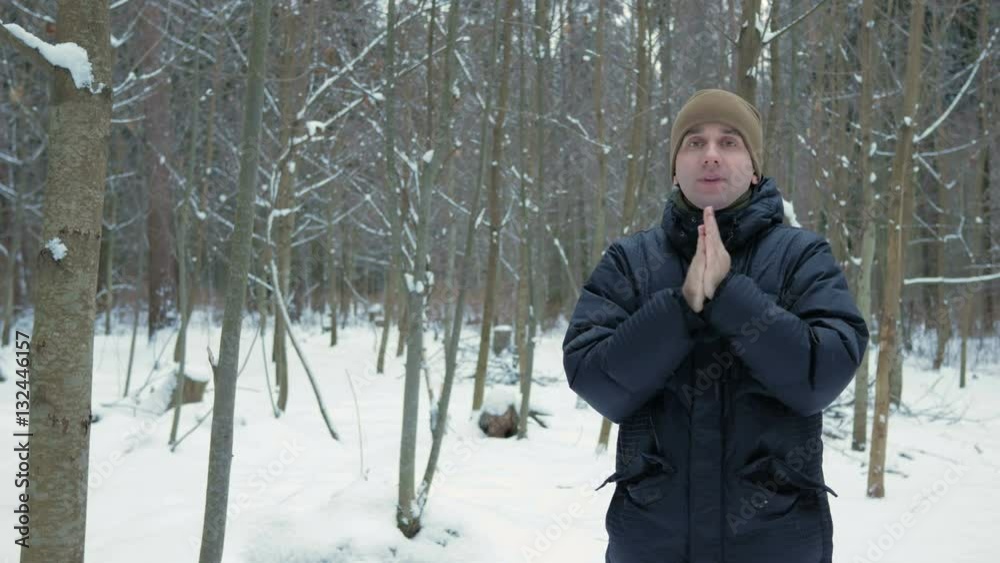 A young man in winter forest freeze. He's breathing on his hands, rubs and wears a hood jackets. Snowy landscape. He looks into the camera A young man in winter forest freeze. He's breathing on his hands, rubs and wears a hood jackets. Snowy landscape. He looks into the camera