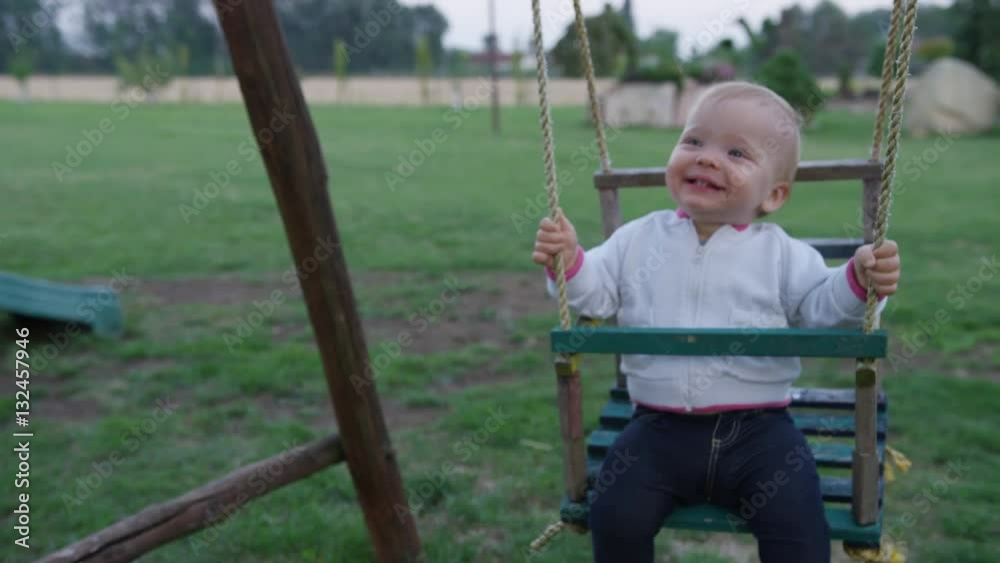 A baby girl happily swings back and forth on a wooden swing set in a garden. At one point she rocks her legs and head back and forth in joy. 4k. Slow motion. A baby girl happily swings back and forth on a wooden swing set in a garden. At one point she rocks her legs and head back and forth in joy. 4k. Slow motion.