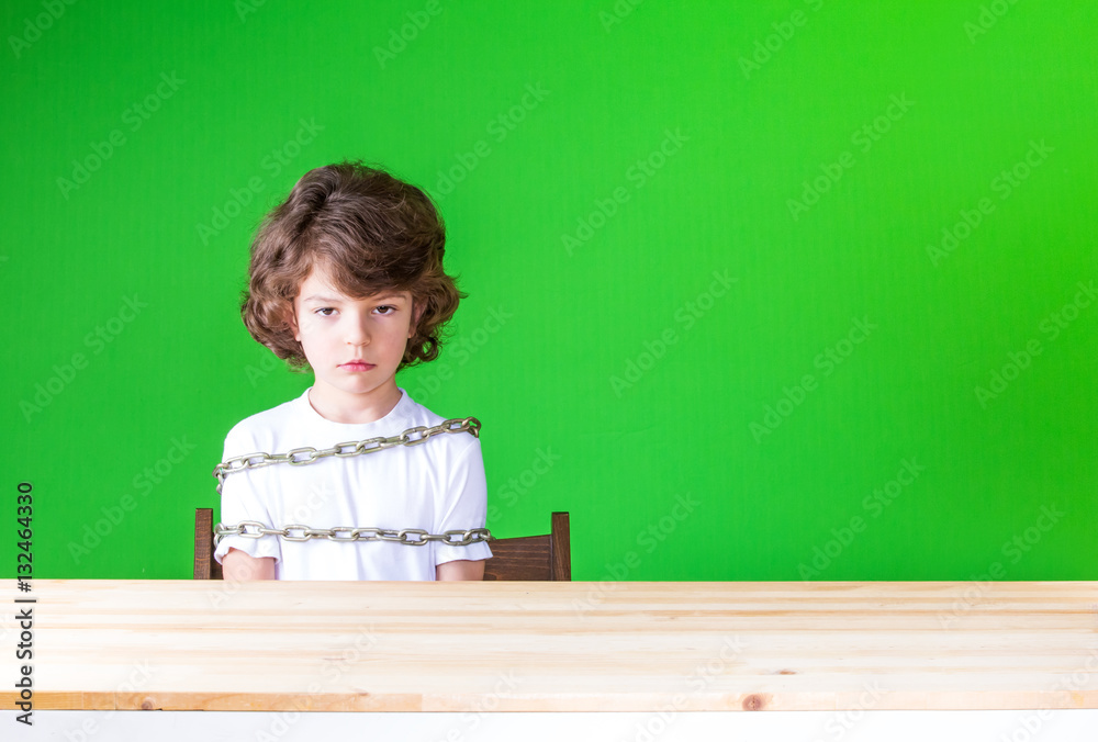 Curly cute boy sitting at the table, wrapped a chain, and sadly looks at the camera. Close-up. Green background. Curly cute boy sitting at the table, wrapped a chain, and sadly looks at the camera. Close-up. Green background.