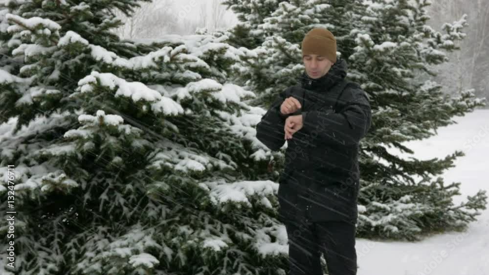 A man checks the messages on the smart watch in the winter snowy forest. Big snowfall. He dictates a voice response to the clock. A man dressed in a dark warm jacket. A man checks the messages on the smart watch in the winter snowy forest. Big snowfall. He dictates a voice response to the clock. A man dressed in a dark warm jacket.