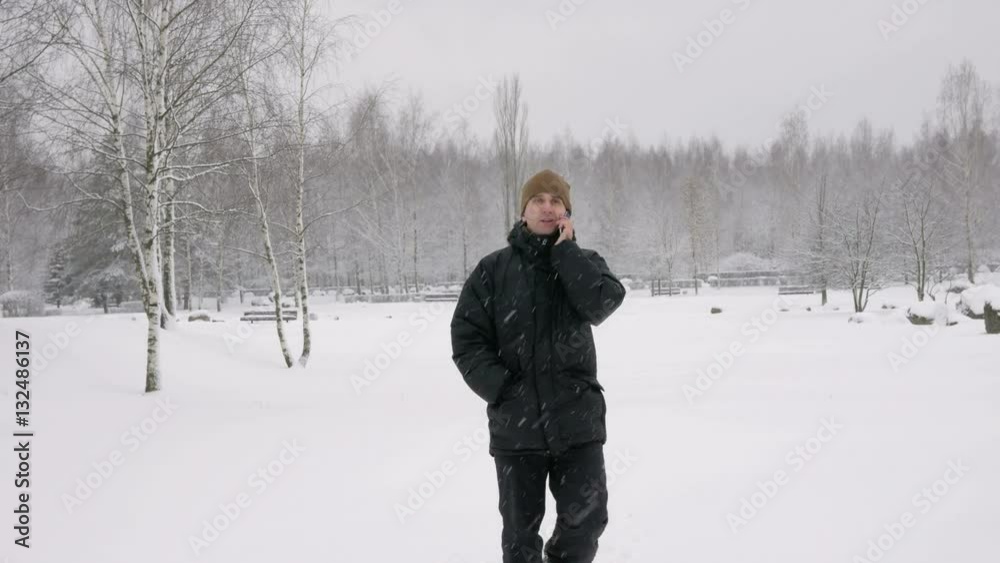 A young man goes on a winter forest and talking on the phone. Big snowfall. He stops and ends the conversation. A man in a dark jacket and a warm hat. A young man goes on a winter forest and talking on the phone. Big snowfall. He stops and ends the conversation. A man in a dark jacket and a warm hat.