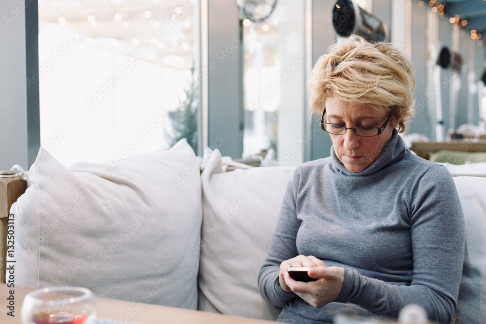 Mid age woman with mobile phone sitting at indoor cafe looking for screen Mid age woman with mobile phone sitting at indoor cafe looking for screen