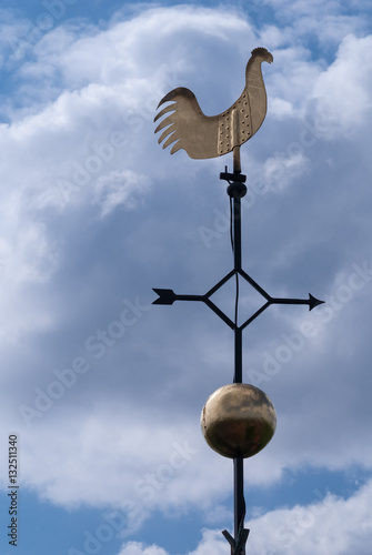 A weather cock against a blue sky with white clouds.