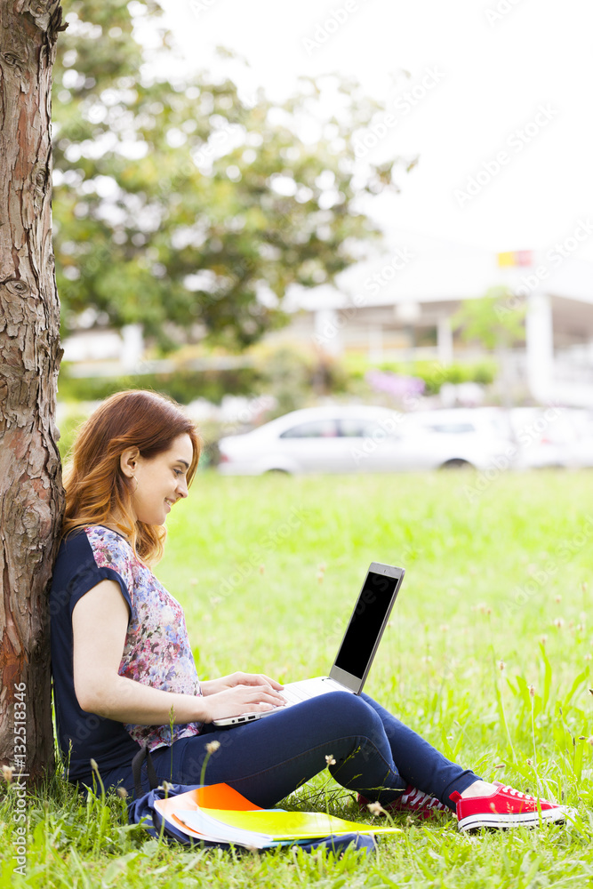 Beautiful student girl sitting on the grass and using laptop com Beautiful student girl sitting on the grass and using laptop com