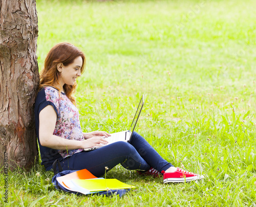 Pretty student girl sitting on the grass and using laptop comput