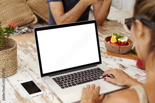 Young fashionable woman wearing sunglasses sitting at wooden table with open notebook computer, food and blank touchscreen mobile phone and enjoying free high-speed internet connection at cafe