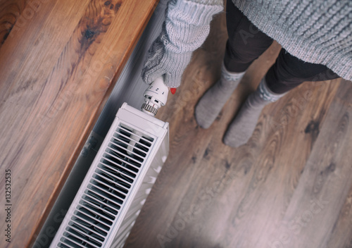 Close up of woman hands wearing woolen gloves turning heater up. Snow at the windowsill. Winter energy saving concept.
