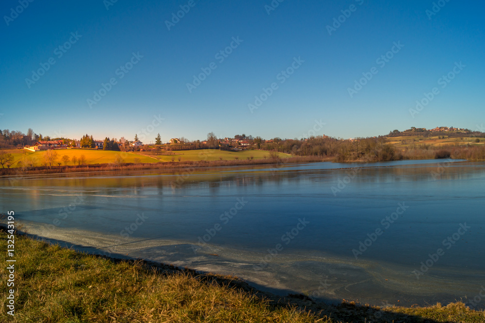 expanse of green grass on the lake with blue sky, countryside and farm around the lake, sunlit water