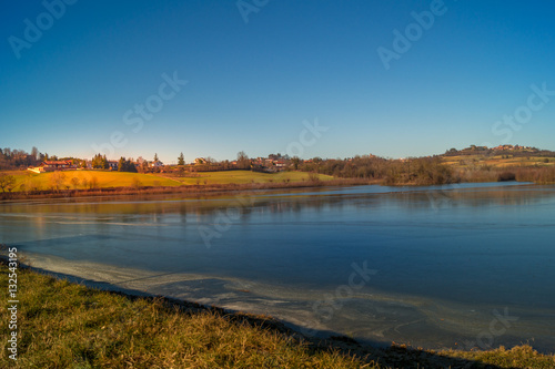 expanse of green grass on the lake with blue sky, countryside and farm around the lake, sunlit water