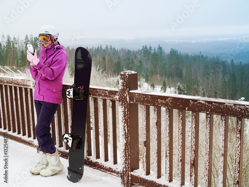 Attractive girl on mountain with snowboard. Snowboarder with mobile phone in hand. Girl snowboard helmet phone suit sunglasses.