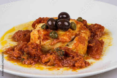 Italian codfish with black olives, cappers and tomato in a design plate, isolated on a white background
