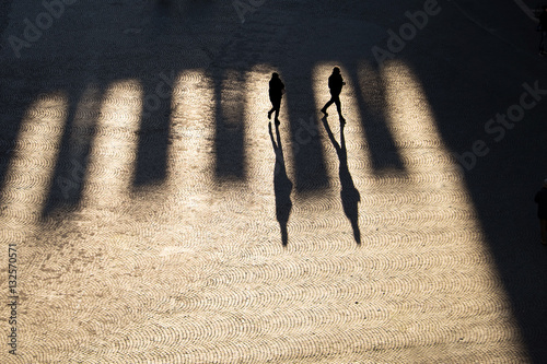 Due persone in controluce con ombre lunghissime camminano in una piazza assolata