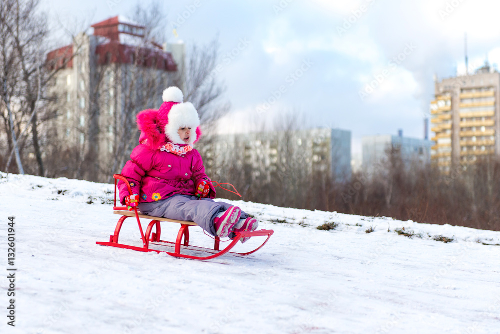 The child, a little girl riding on a sled with snow slides. Winter fun for children.