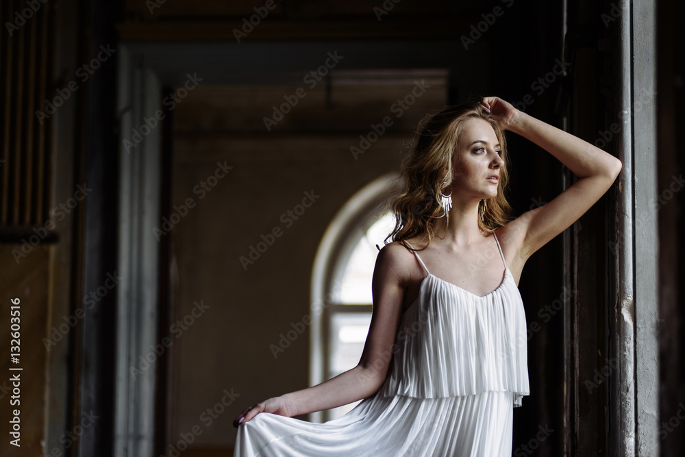 Indoor summer portrait of young pretty cute girl. Beautiful woman posing beside fairytale window inside wood cabinet, scars old castle. Bride in white atlas dress. Photo has a model.