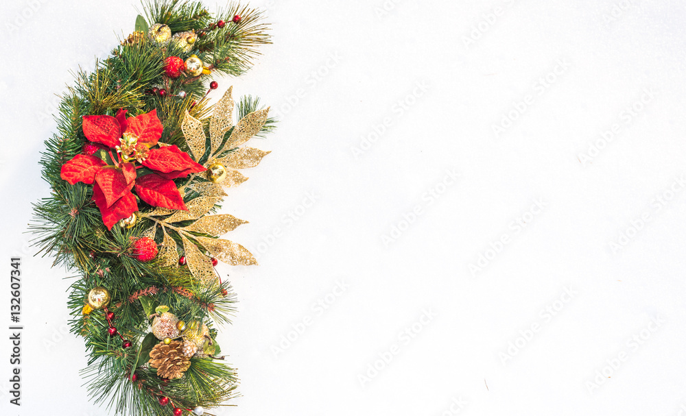 Christmas holiday faux poinsettia pine wreath adorned with gold leaf ribbon, Christmas balls and holly, resting on natural snow with white copy space.