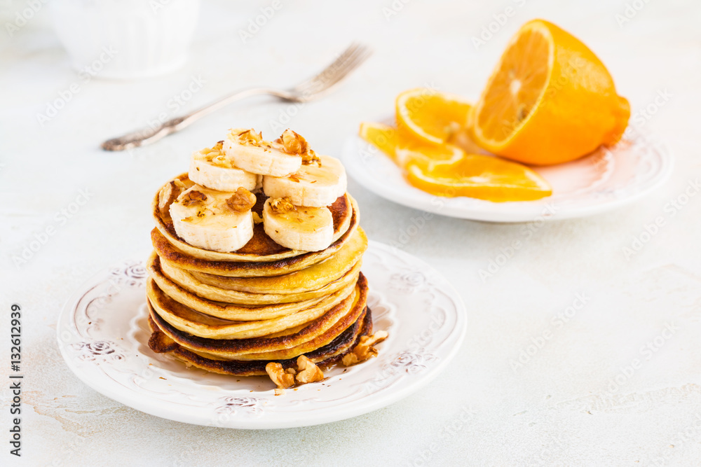 Stack of homemade pancakes with banana, maple syrup and walnuts on vintage plate. Fork, fresh sliced lemon, white and gray concrete background.