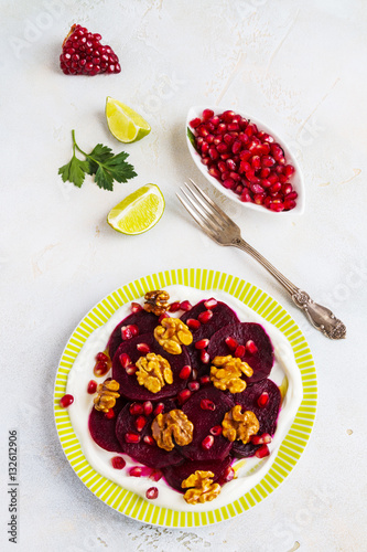 Dietary vegetarian salad of roasted beets with pomegranate seeds, walnuts caramelized in honey and natural yoghurt. Slices of lime, vintage fork, white and gray table, top view.