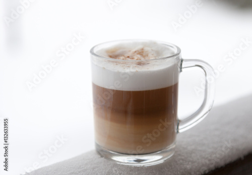 Cup with cappuccino coffee on the wooden bench covered with snow. Blurred winter background with snowflakes. 
